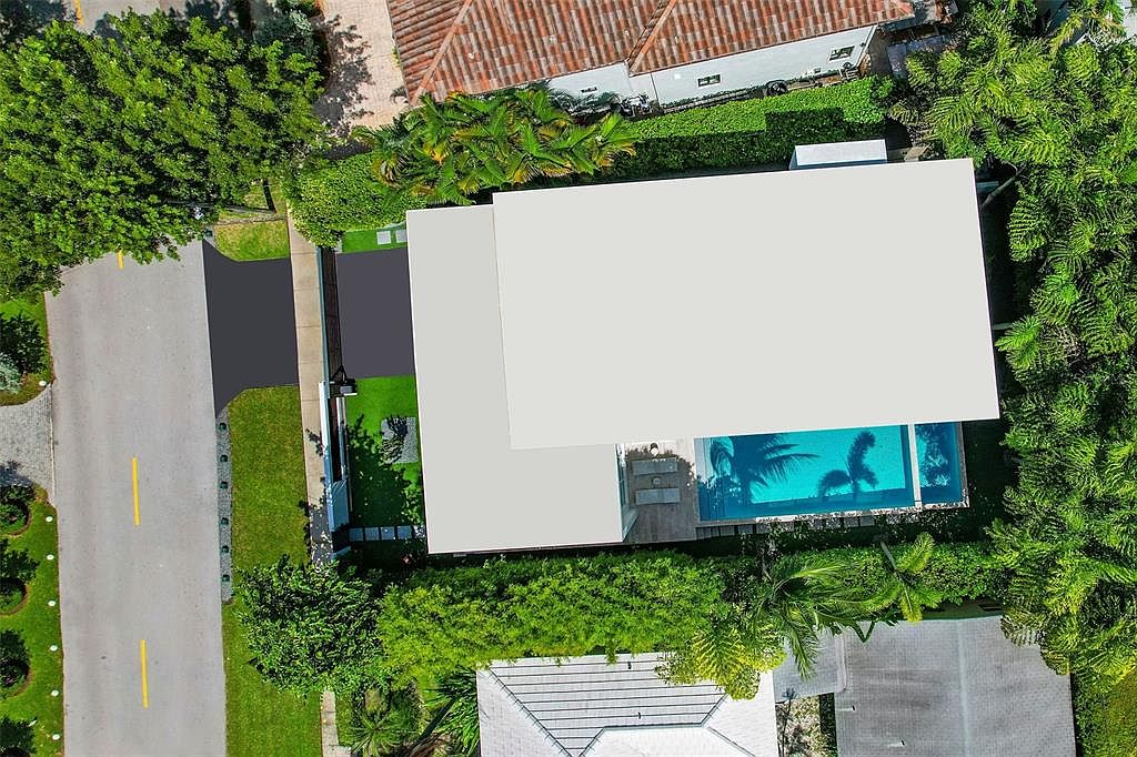 This high-angle aerial view captures a modern residential property featuring a flat-roofed home with a private swimming pool. The property is surrounded by lush green landscaping and mature trees, providing a sense of privacy and tranquility. The layout includes a paved driveway leading to the entrance, showcasing a clean, contemporary architectural style.