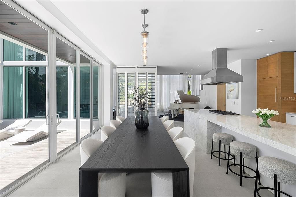 This sophisticated open-concept dining area features a long, dark wood table surrounded by modern white upholstered chairs, positioned adjacent to a sleek marble-topped kitchen island. Large floor-to-ceiling glass sliding doors provide an abundance of natural light and a seamless transition to the outdoor patio, while a grand piano is visible in the background. The space exudes a minimalist, high-end contemporary aesthetic with clean lines and a neutral color palette.