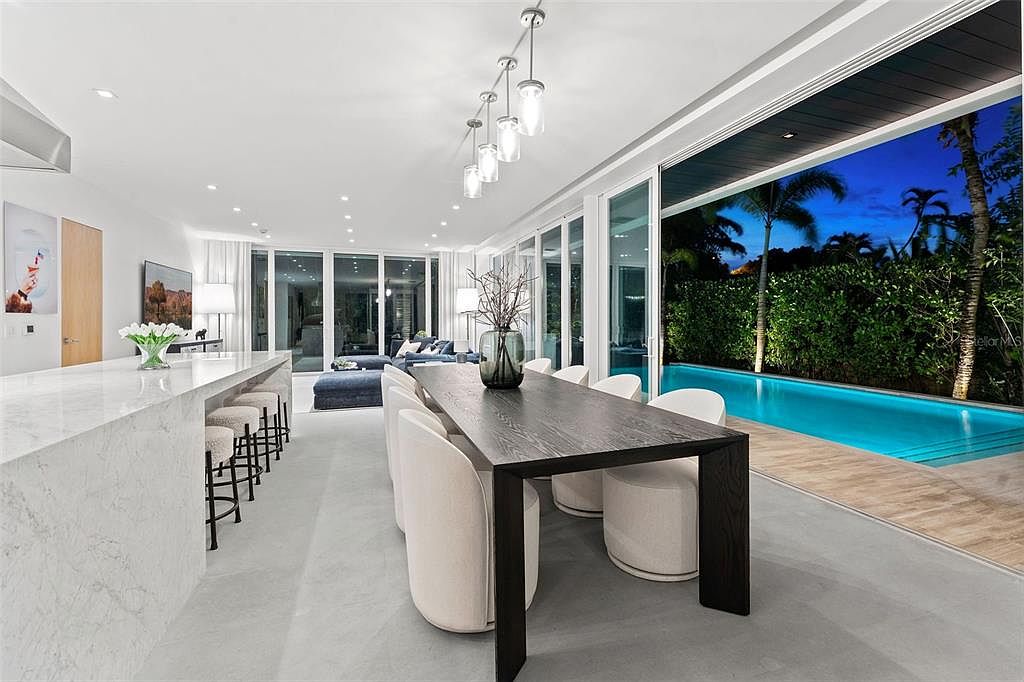 This sophisticated open-concept dining area features a sleek, dark wood table paired with modern, cream-colored upholstered chairs, creating a striking contrast against the minimalist white interior. The space seamlessly transitions to an outdoor pool area through expansive floor-to-ceiling glass sliding doors, enhancing the indoor-outdoor living experience. A contemporary linear pendant light fixture hangs above the table, while a marble-topped kitchen island with bar seating completes this luxurious, high-end living space.
