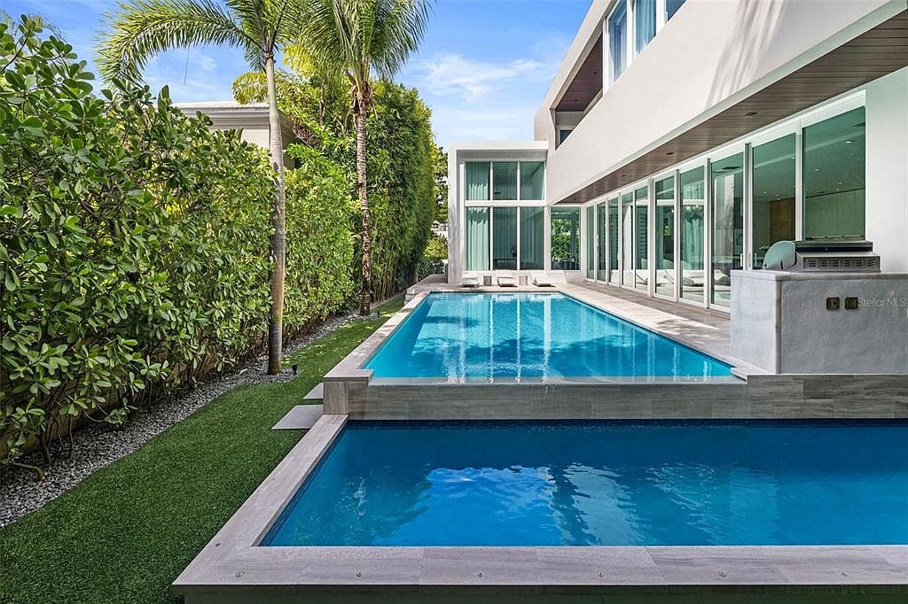 This modern, multi-level swimming pool area features a sleek, geometric design with a raised upper pool cascading into a lower pool. The contemporary white architecture of the home, characterized by expansive floor-to-ceiling glass walls, creates a seamless indoor-outdoor transition. Lush green landscaping and tall palm trees frame the space, offering a private and luxurious resort-like atmosphere.