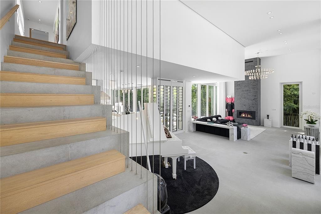 This expansive, modern living area features a striking staircase with concrete steps and wooden treads, separated by a sleek cable railing. The open-concept space includes a white grand piano, a contemporary fireplace with a dark stone surround, and a bold black-and-white aesthetic. Large windows and high ceilings create an airy, sophisticated atmosphere, perfect for luxury entertaining.