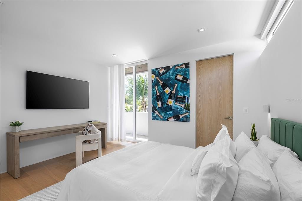 This modern primary bedroom features a minimalist aesthetic with crisp white walls, light wood flooring, and a large bed with a green upholstered headboard. A sleek wooden desk with a matching chair sits beneath a wall-mounted television, while a vibrant piece of art depicting champagne bottles adds a playful touch of color. The room is bright and airy, with a sliding glass door leading to a balcony that brings in natural light and a view of palm trees.