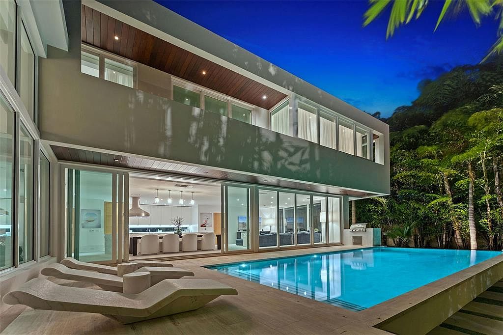 This stunning modern residence features a seamless indoor-outdoor transition with expansive floor-to-ceiling glass walls that open onto a sleek, illuminated swimming pool. The patio area is furnished with contemporary lounge chairs, creating a luxurious resort-like atmosphere perfect for evening relaxation. The architectural design emphasizes clean lines, warm wood accents on the overhang, and a sophisticated, high-end aesthetic.