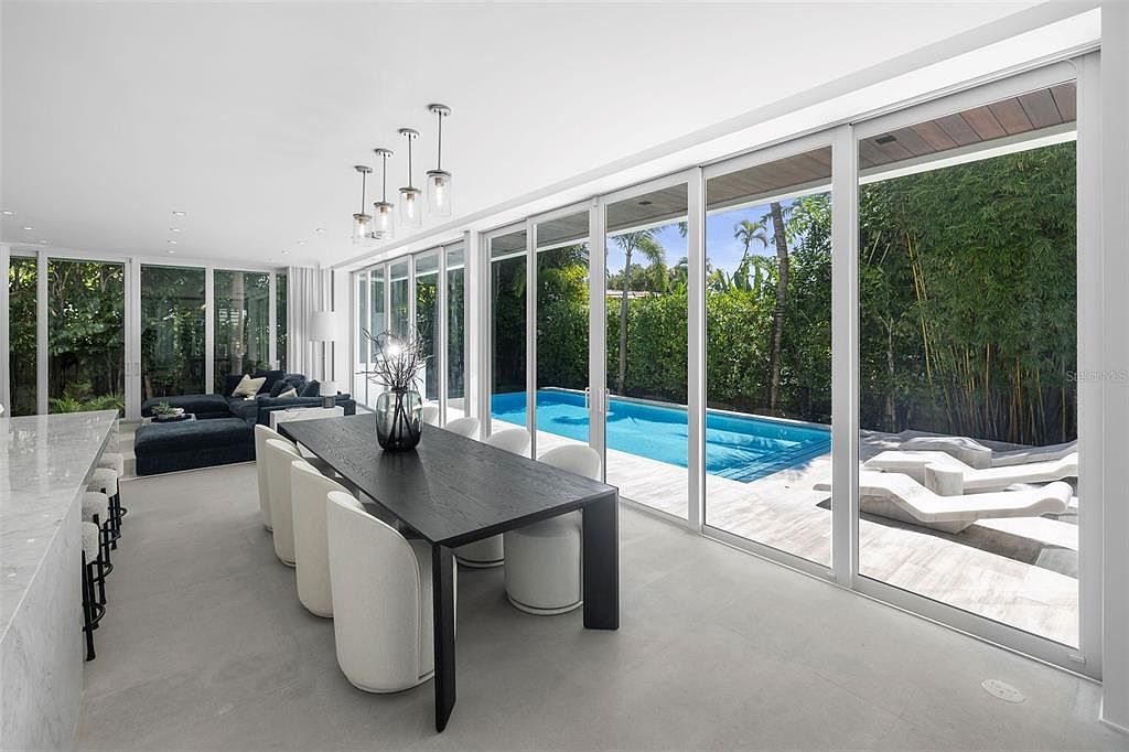 This modern, open-concept dining area features a sleek black table surrounded by minimalist white chairs, set against a backdrop of floor-to-ceiling glass sliding doors. The space seamlessly connects to an outdoor pool area, creating a bright and airy atmosphere with a contemporary aesthetic. The neutral color palette and clean lines emphasize a sophisticated, indoor-outdoor living experience.