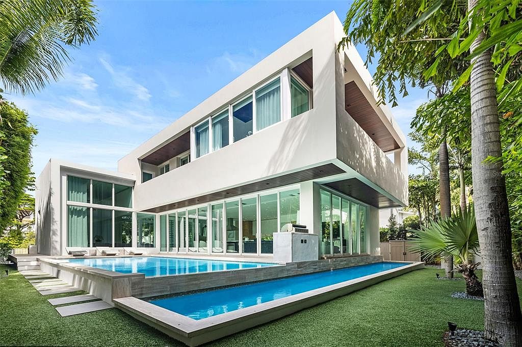 This modern, architectural luxury home features a striking two-story design with clean lines, expansive floor-to-ceiling glass walls, and dark wood accents. The property boasts a sophisticated multi-level pool area, including a main swimming pool and a long, narrow lap pool set against a lush, manicured lawn. The perspective captures the seamless integration between the indoor living spaces and the outdoor oasis, emphasizing a high-end, resort-style lifestyle.