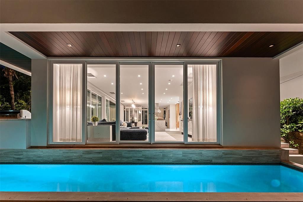 This image captures a serene evening view of a modern home's backyard, featuring a vibrant, illuminated swimming pool directly adjacent to a large glass sliding door entrance. The architectural design highlights a sleek, dark wood-paneled ceiling overhang that frames the interior living space, which is visible through the glass. The combination of the glowing pool water and the warm interior lighting creates an inviting, luxurious atmosphere perfect for evening relaxation.