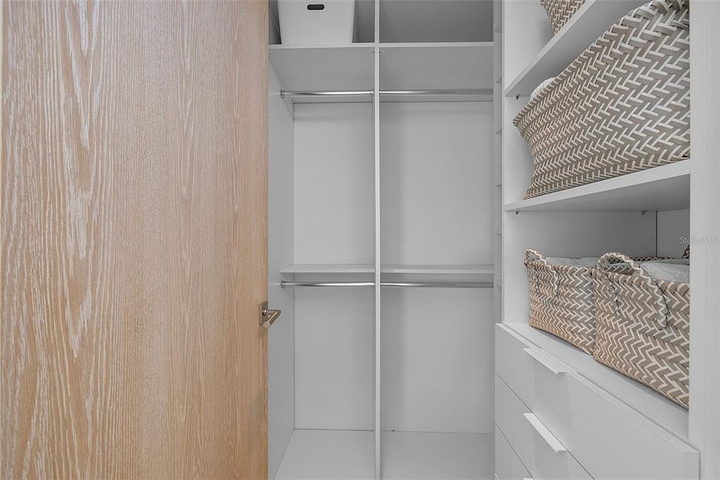 This organized walk-in closet features a clean, modern aesthetic with white shelving, hanging rods, and built-in drawers. The space is accented by woven storage baskets on the open shelves and a light wood-toned door, creating a bright and functional storage area. The perspective is a straight-on view looking into the closet, highlighting its efficient use of space.
