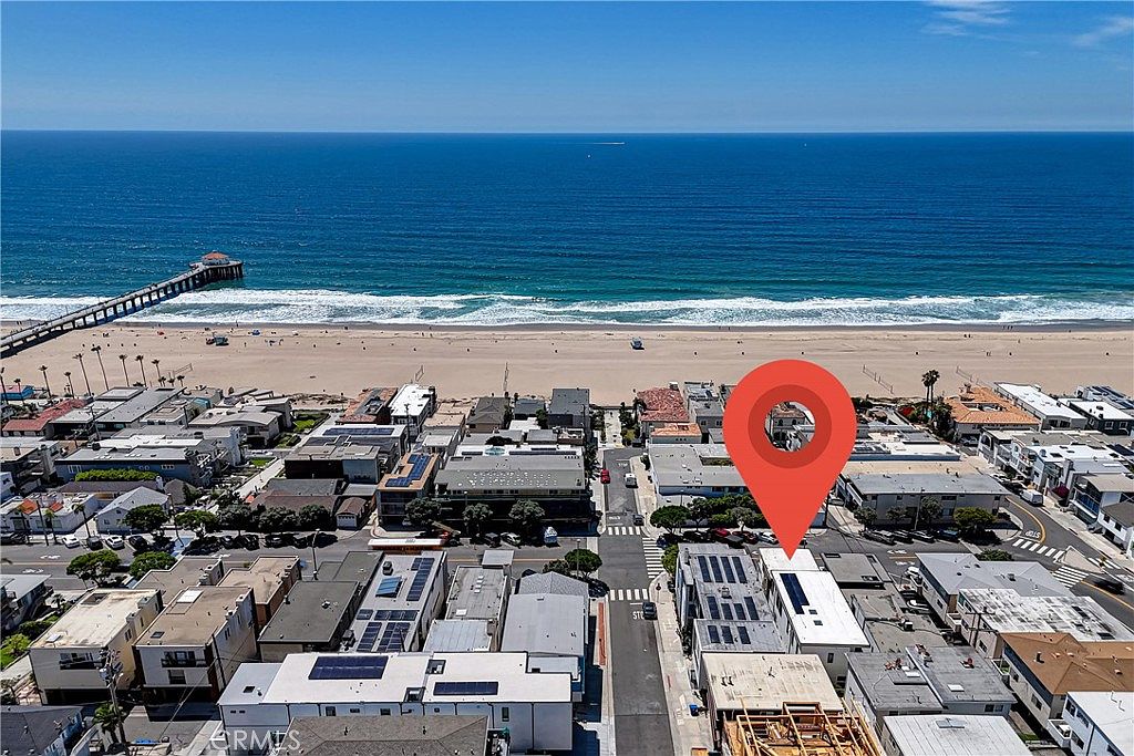This high-angle aerial view captures a coastal neighborhood situated directly along a sandy beach and the Pacific Ocean. A prominent red location pin highlights a specific property within the dense residential grid, which features flat-roofed buildings and proximity to a public pier. The perspective emphasizes the prime beachside location and the expansive ocean horizon under a clear blue sky.