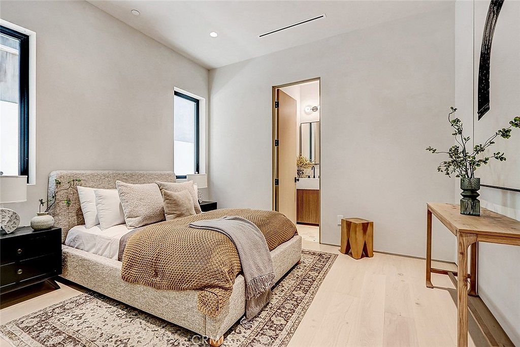 This modern bedroom features a neutral color palette with light wood flooring and soft gray walls. A plush bed with a textured throw blanket serves as the focal point, flanked by a dark nightstand and a minimalist wooden console table. A doorway leads into an adjacent bathroom, creating a seamless flow between the sleeping and personal care spaces.