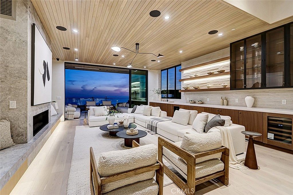 This expansive, modern living room features a light wood-paneled ceiling and a large, open-concept layout that flows seamlessly toward a balcony with ocean views. The space is anchored by a large white sectional sofa, two textured armchairs, and a stone-clad chimney breast housing a fireplace. A built-in bar area with illuminated shelving and glass-front cabinetry adds a sophisticated touch, while the neutral color palette creates a serene and luxurious atmosphere.