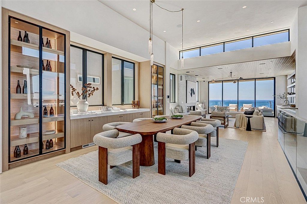 This sophisticated dining area features a long, warm-toned wooden table surrounded by six upholstered chairs, set atop a textured area rug. To the left, a built-in display cabinet with glass doors and integrated lighting provides elegant storage, while the open-concept layout flows seamlessly into a bright living space with expansive glass walls overlooking the ocean. The room is defined by its high ceilings, minimalist aesthetic, and a neutral color palette that emphasizes the natural light and coastal views.