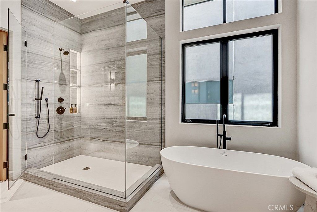 This modern primary bathroom features a large walk-in shower with a glass enclosure and light-toned, horizontally-veined stone wall tiles. Adjacent to the shower is a sleek, freestanding white soaking tub positioned beneath a large window that allows for ample natural light. The space is finished with dark, matte-black plumbing fixtures, creating a sophisticated and high-end aesthetic.