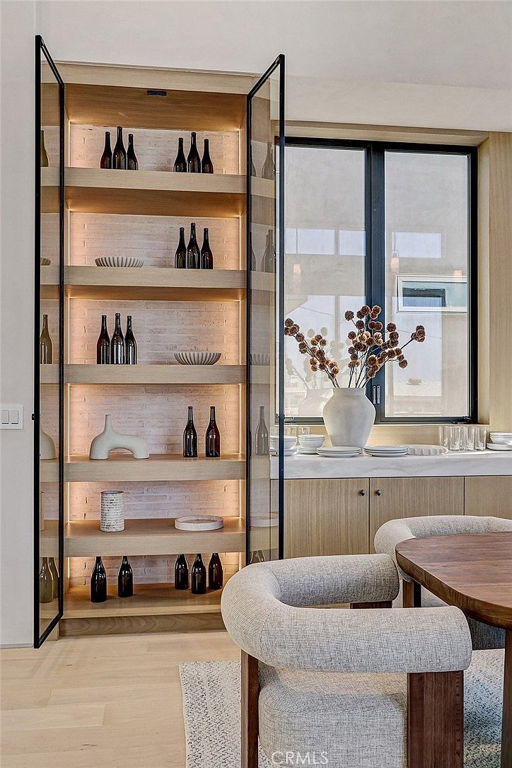 This dining area features a sophisticated built-in display cabinet with glass doors, illuminated shelving, and a textured brick back wall. A wooden dining table is paired with curved, upholstered chairs, while a large window provides natural light and a view of the outdoors. The space is accented by a decorative vase with dried botanicals, creating a warm and modern aesthetic.