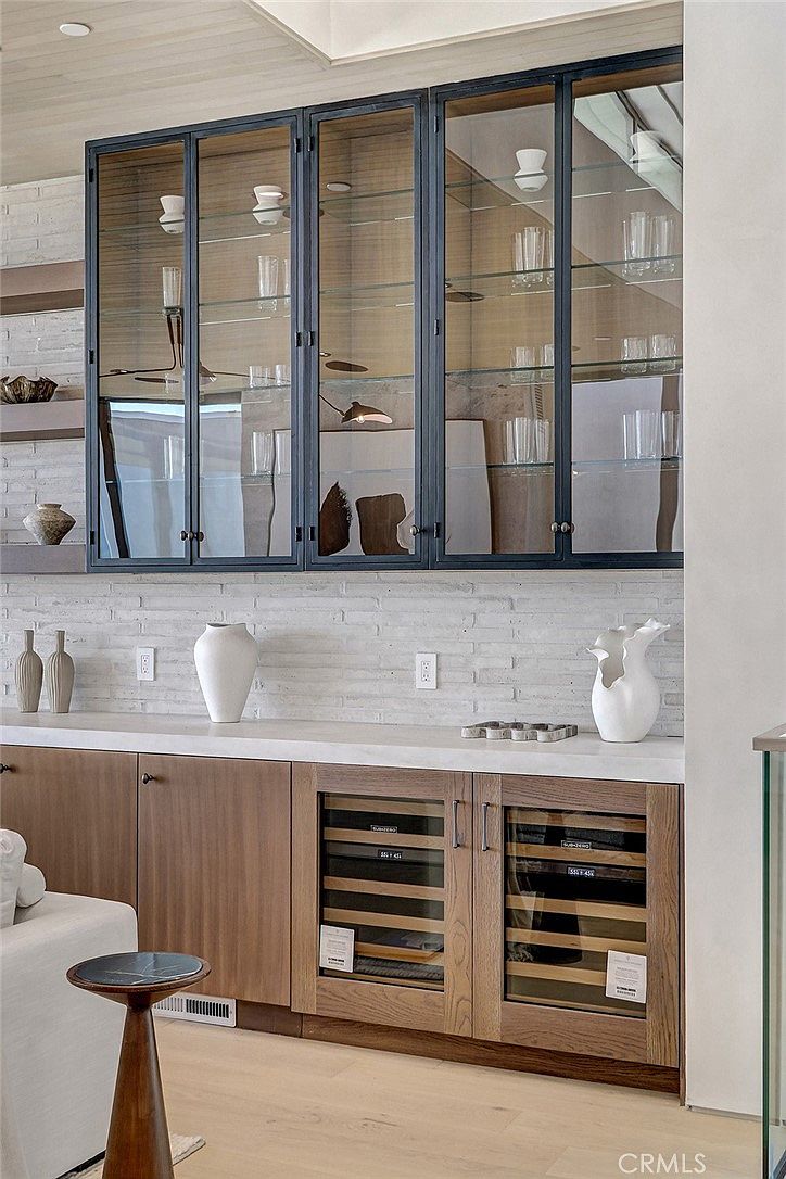 This sophisticated wet bar or butler's pantry area features custom wood cabinetry paired with a light-toned stone countertop and a textured white brick backsplash. Above the counter, sleek black-framed glass cabinets provide elegant display storage for glassware, while the lower section includes integrated wine refrigeration units. The space is accented by minimalist decor, including sculptural vases and open shelving, creating a refined and functional entertaining area.