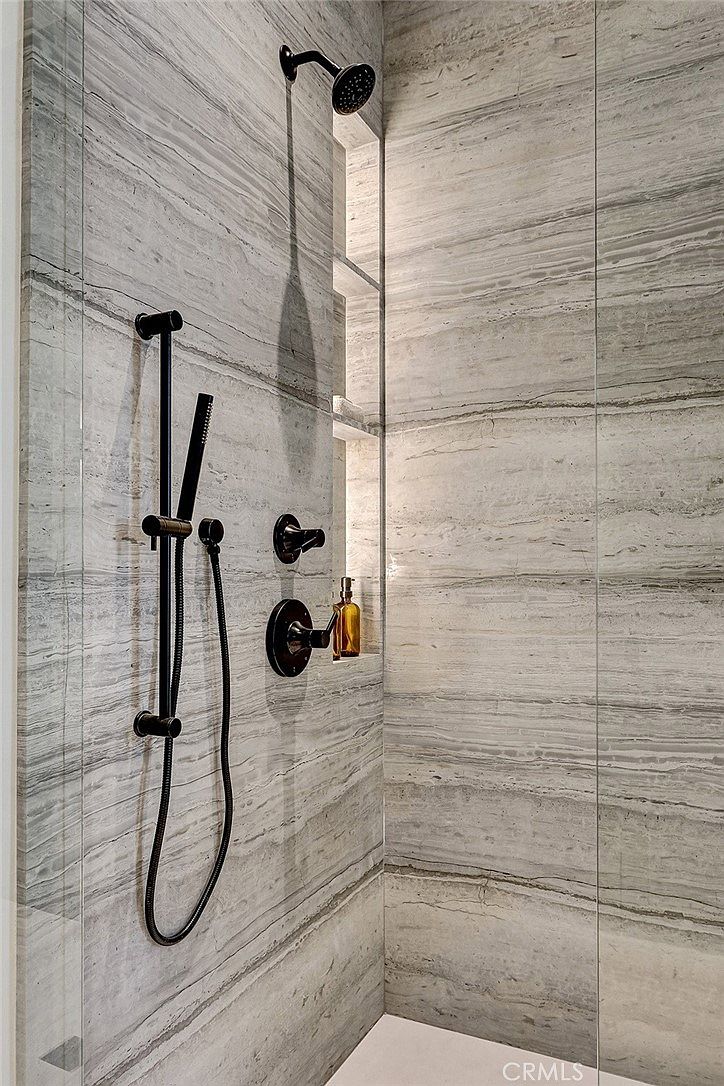 This modern shower features large-format, light-toned stone wall tiles with prominent horizontal veining. The space is equipped with matte black fixtures, including a rainfall showerhead, a handheld sprayer on a slide bar, and integrated controls. A recessed, illuminated niche provides convenient storage for toiletries, adding a functional and sophisticated touch to the minimalist design.