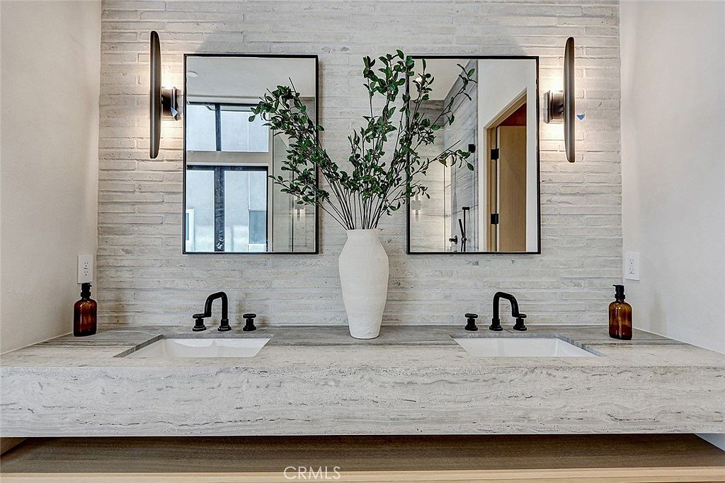 This modern double vanity features a long, thick stone countertop with integrated sinks and matte black fixtures. The wall behind the vanity is finished with a textured, light-colored stone tile, complemented by two rectangular mirrors and sleek vertical wall sconces. A central vase with greenery adds a natural touch to the clean, minimalist aesthetic of the space.