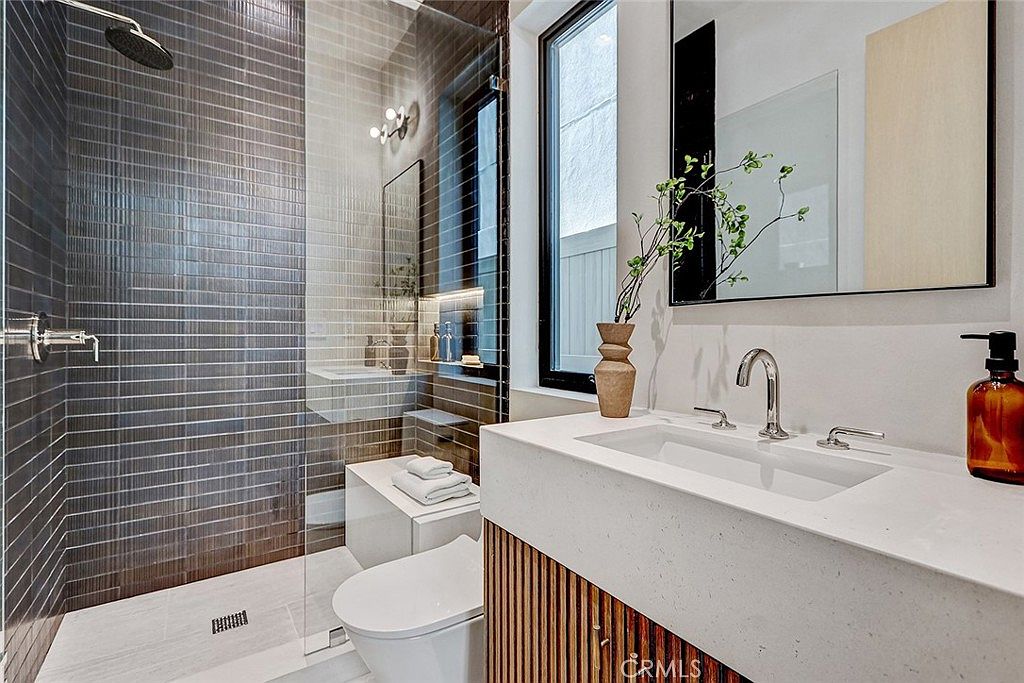 This modern bathroom features a walk-in shower with dark, horizontally stacked tiles and a glass partition. A vanity with a light-colored stone countertop and a slatted wood base sits beside a white toilet, while a tall window provides natural light. The space is accented with a decorative vase and a mirror, creating a clean and contemporary aesthetic.
