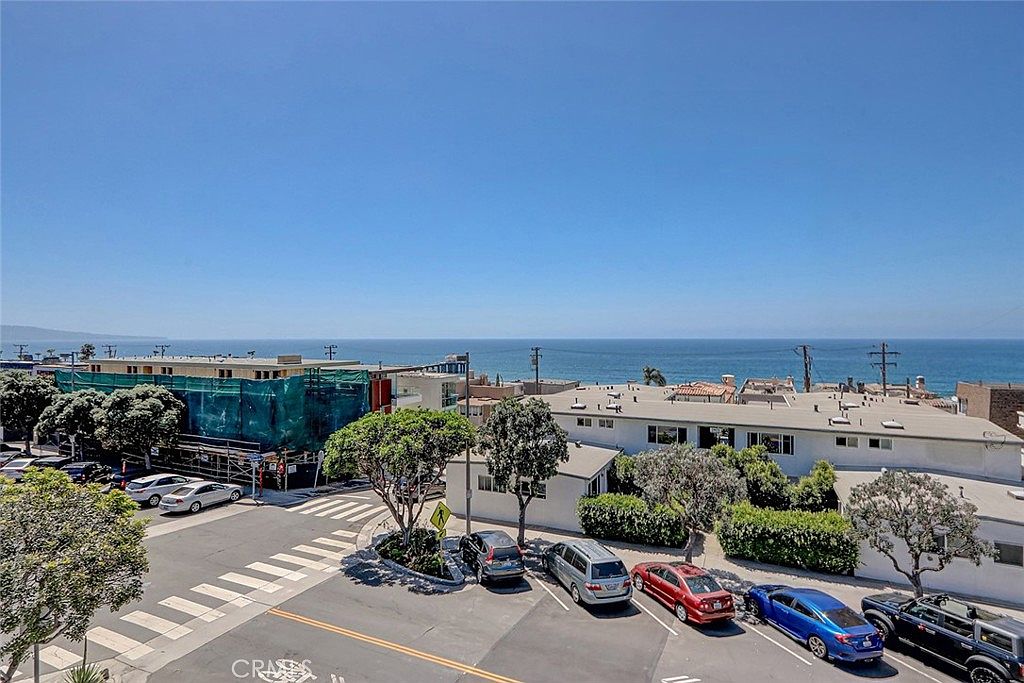 This high-angle aerial view captures a coastal neighborhood street scene with a clear, expansive view of the ocean horizon. The foreground features a paved parking area with several parked cars, while the mid-ground shows a mix of residential buildings, including one undergoing construction with green protective netting. The bright, sunny day highlights the proximity of the property to the coastline, emphasizing a desirable beachside location.