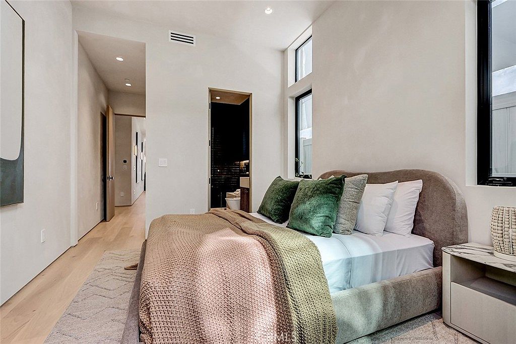 This modern bedroom features a neutral color palette with light wood flooring and soft, textured bedding. A plush, upholstered bed frame serves as the focal point, accented by green throw pillows and a draped knit blanket. Tall windows allow natural light to fill the space, while a doorway leads into a hallway and an adjacent bathroom.