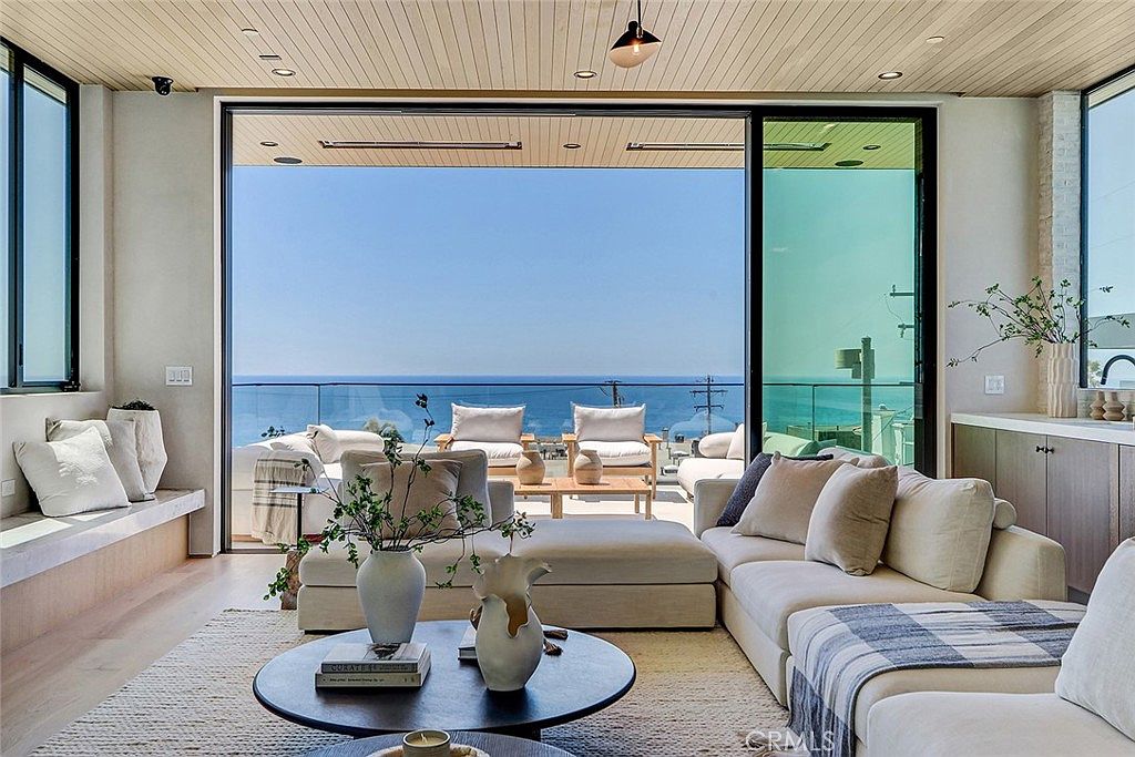 This bright and airy living room features a seamless transition to an outdoor deck through expansive floor-to-ceiling glass sliding doors that frame a panoramic ocean view. The interior is furnished with a large, light-toned sectional sofa, a circular coffee table, and a built-in bench seating area along the side wall. The space is finished with light wood flooring and a slatted wood ceiling, creating a cohesive, modern coastal aesthetic.