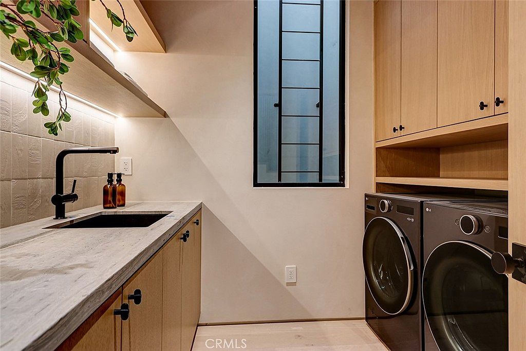 This modern laundry room features light wood cabinetry, a stone countertop with an integrated sink, and a matte black faucet. A pair of dark-toned laundry appliances is positioned under a built-in shelf, while a tall, narrow window with a black frame provides natural light. The space is finished with neutral wall tones and light-colored flooring, creating a clean and functional aesthetic.
