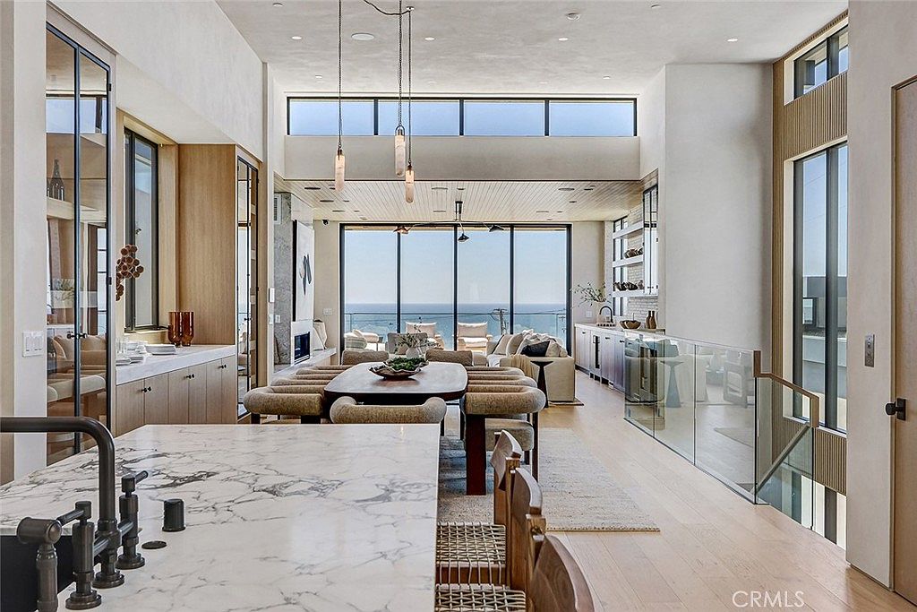 This expansive, open-concept living space features a prominent kitchen island with a stone countertop in the foreground, leading into a dining area and a living room with panoramic ocean views. The room is defined by high ceilings, light wood flooring, and a wall of large glass doors that seamlessly connect the interior to the outdoor scenery. The design aesthetic is modern and airy, characterized by clean lines, neutral tones, and sophisticated pendant lighting.
