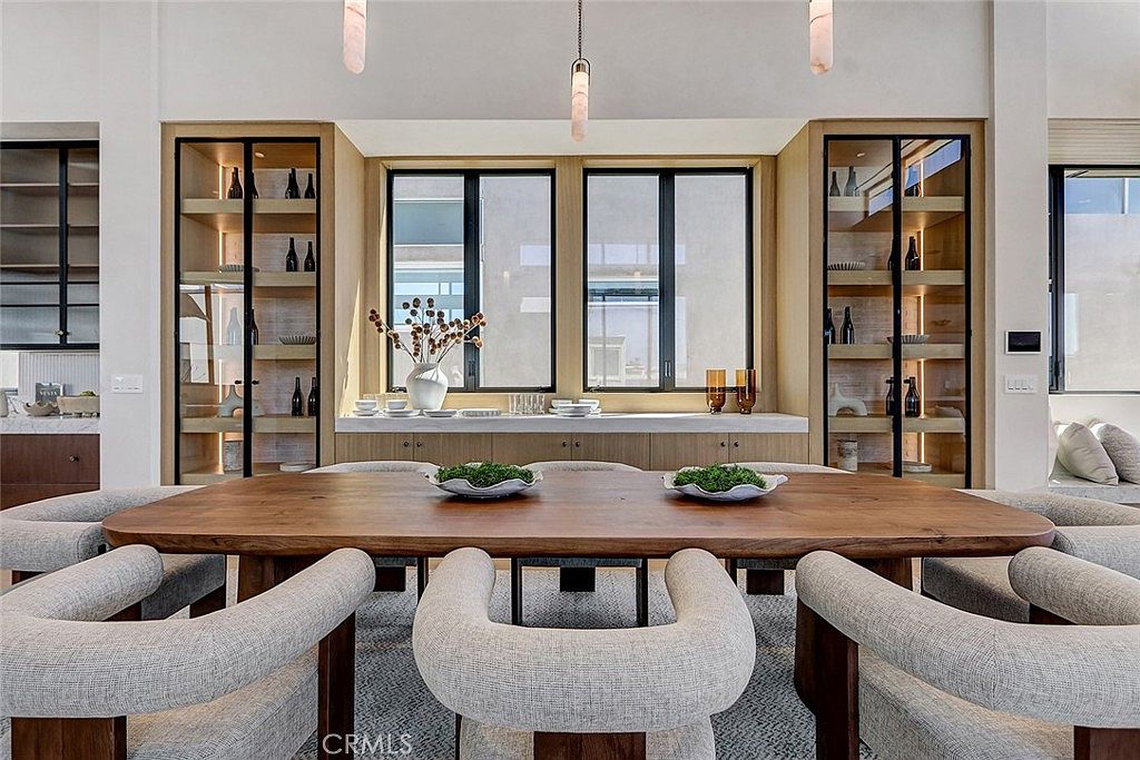 This elegant dining room features a large, organic-shaped wooden table surrounded by six upholstered, curved-back chairs. The space is anchored by a built-in light wood cabinetry unit with glass-front display shelves and integrated lighting, framing a central window area. The room is illuminated by three modern, slender pendant lights, creating a sophisticated and inviting atmosphere.