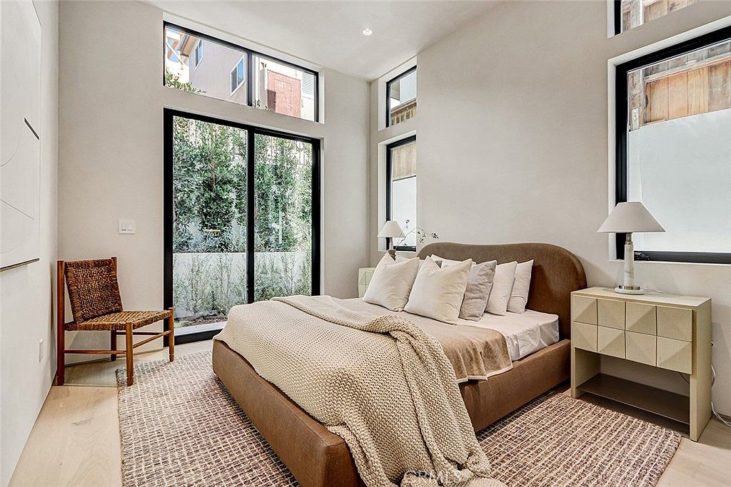 This modern bedroom features a low-profile bed with a curved headboard, dressed in neutral linens and a textured throw blanket. The room is illuminated by natural light from large windows and a sliding glass door that opens to a private outdoor area. A woven chair and a light-toned bedside table with a lamp complete the minimalist, serene aesthetic.