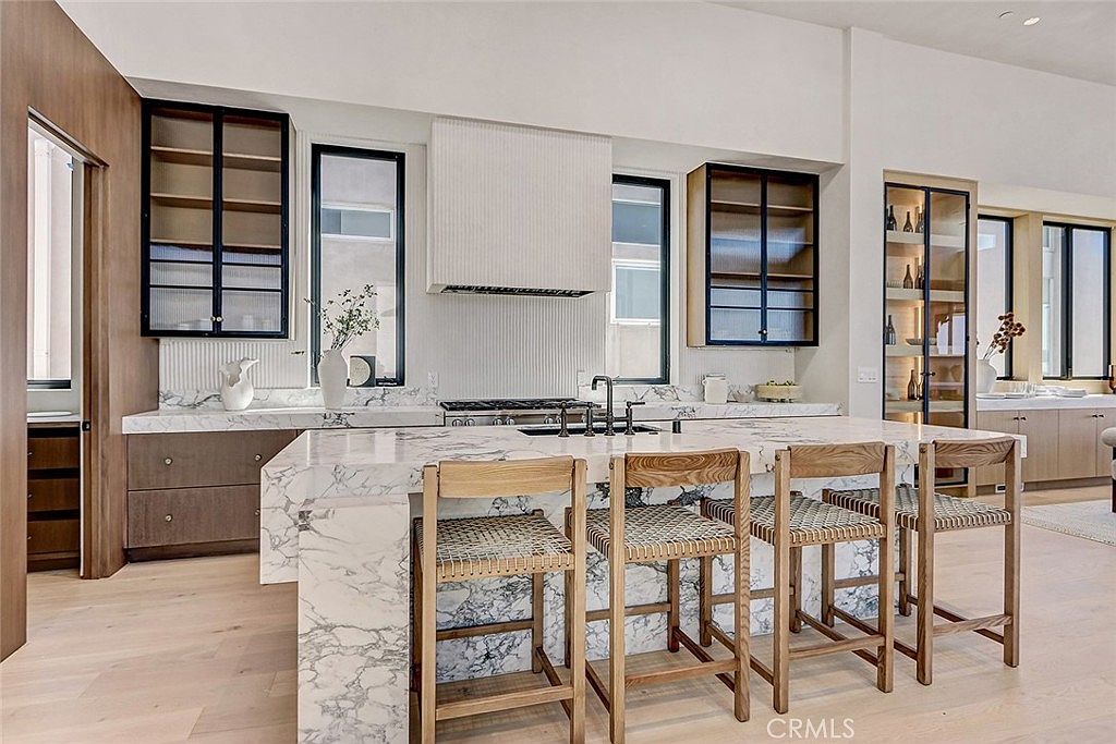 This modern kitchen features a large central island with a stone waterfall countertop and four woven-seat bar stools. The space is anchored by a custom range hood and a professional-style gas cooktop, flanked by glass-front cabinetry and tall windows that bring in natural light. The design utilizes a neutral palette with light wood flooring and stone surfaces, creating a sophisticated and airy atmosphere.