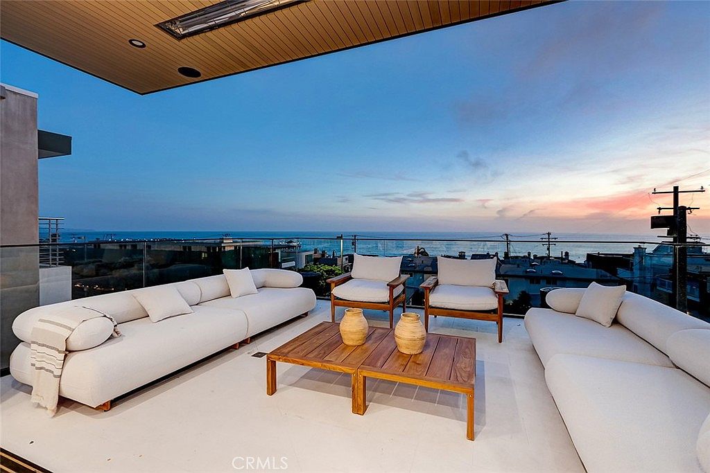 This expansive, modern balcony offers a stunning panoramic view of the ocean at sunset. The space is furnished with two large, light-colored modular sofas and a pair of wooden armchairs arranged around a central wooden coffee table with decorative vases. A glass railing ensures an unobstructed view, while a covered ceiling section with integrated lighting provides a sophisticated outdoor living experience.