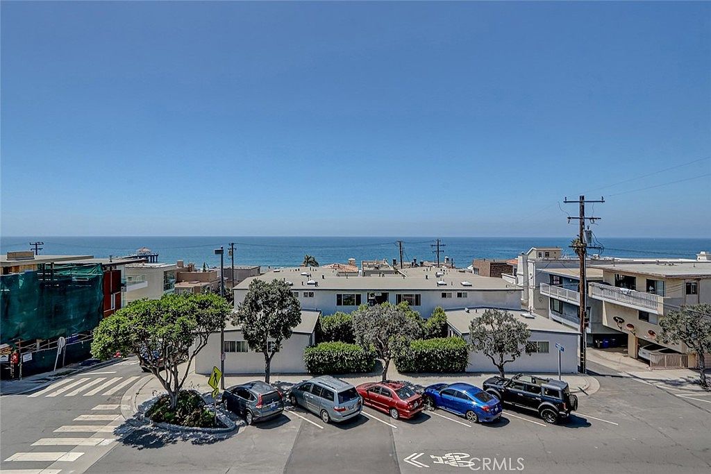 This elevated, wide-angle shot captures a multi-unit residential building situated along a coastal street, offering a clear view of the ocean horizon. The scene features a paved parking area in the foreground with several parked vehicles, mature trees, and a crosswalk leading toward the property. The perspective provides a comprehensive look at the building's exterior, its proximity to the beach, and the surrounding neighborhood layout.
