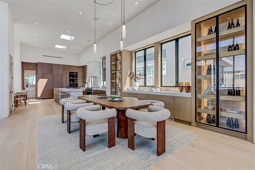 This spacious dining area features a large wooden table surrounded by six upholstered chairs with dark wood frames, set upon a textured area rug. To the right, a tall, glass-fronted display cabinet with integrated lighting showcases glassware, while a long, low console with a stone top sits beneath a row of windows. The open-concept layout flows into a kitchen area in the background, characterized by light wood flooring and high ceilings with recessed lighting.