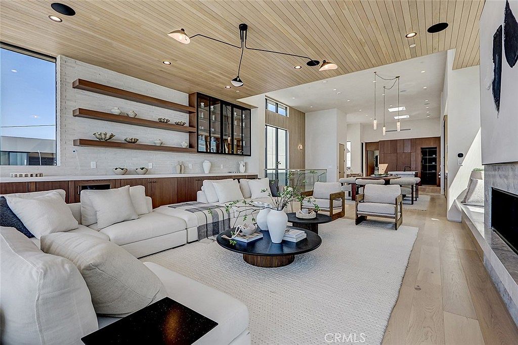 This expansive, open-concept living room features a light wood-paneled ceiling and light-toned hardwood flooring, creating a bright and airy atmosphere. The space is anchored by a large, plush white sectional sofa and a pair of round coffee tables, with a dining area visible in the background. A long, low-profile storage unit with open shelving provides a clean, modern aesthetic, while contemporary light fixtures add a sophisticated touch to the room.