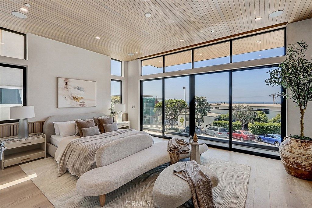 This spacious primary bedroom features a contemporary design with light wood flooring and a slatted wood ceiling. A large bed with neutral-toned bedding is positioned in front of a piece of abstract art, while floor-to-ceiling glass walls provide expansive views of the neighborhood and ocean beyond. The room is furnished with a curved bench at the foot of the bed, matching nightstands, and a large decorative planter, creating a serene and luxurious atmosphere.