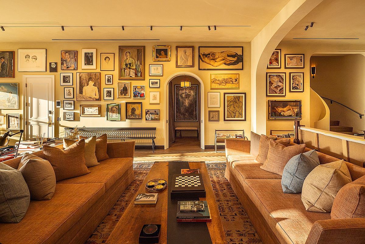 This is an interior shot of a living room featuring two long sofas facing each other, separated by a wooden coffee table. The walls are adorned with numerous framed artworks, creating a gallery-like atmosphere. An arched doorway leads to another room, and a staircase is visible in the background, suggesting a multi-level home.