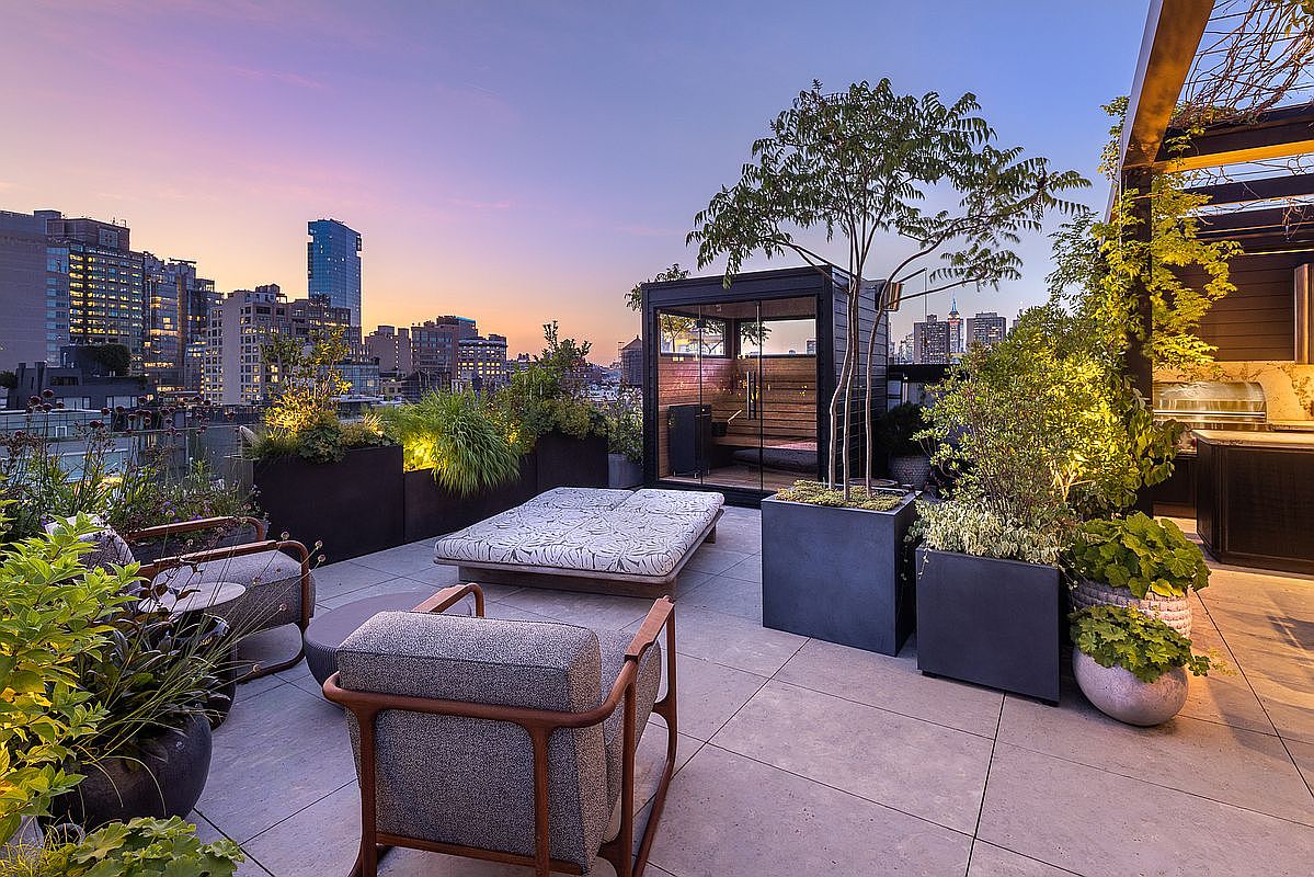 This image showcases a luxurious rooftop patio with stunning city views at dusk. The space is furnished with modern outdoor seating, a daybed, and lush greenery in planters, creating a serene urban oasis. A sauna and outdoor kitchen area are also visible, enhancing the property's appeal for upscale living and entertaining.