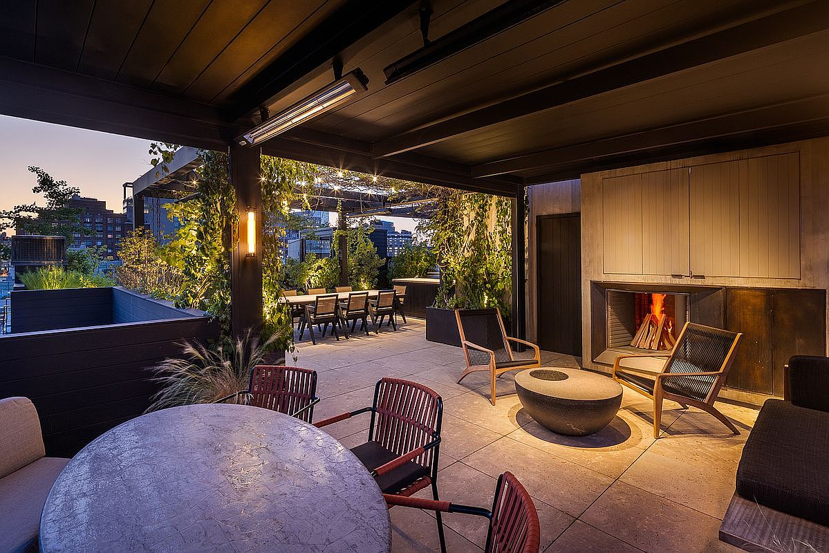 This image showcases a luxurious outdoor patio area, featuring a fireplace, comfortable seating, and a dining table set for several guests. Lush greenery and string lights create an inviting and sophisticated ambiance, perfect for entertaining or relaxing. The space is well-lit and appears to be part of a high-end property with city views.