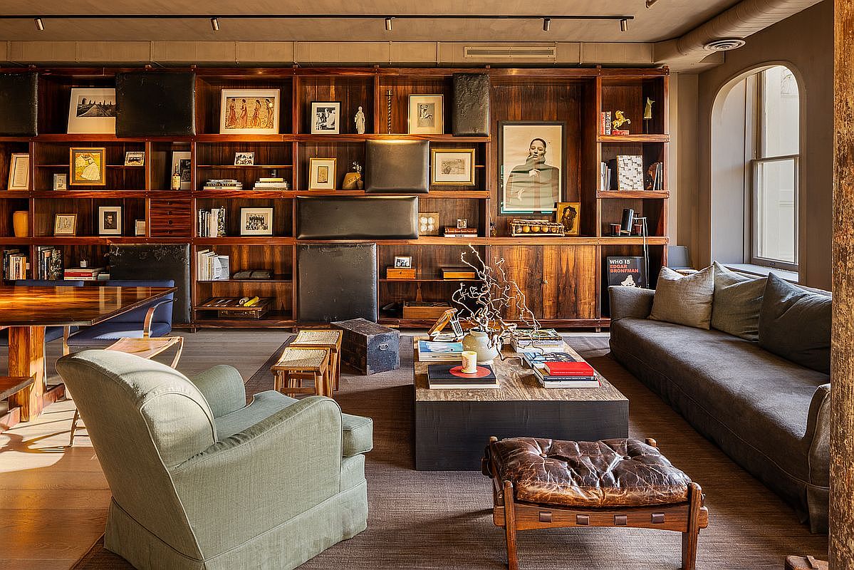 This is an interior shot of a living room featuring a large, dark wood bookshelf filled with books and decorative items. A comfortable gray sofa sits to the right, complemented by a light green armchair and a leather ottoman. The room is well-lit, creating a warm and inviting atmosphere, perfect for relaxation and entertaining.