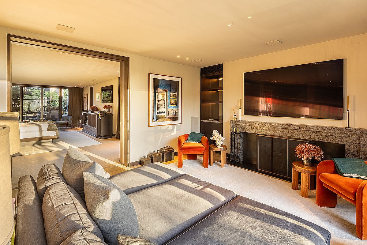 This is an interior shot of a luxurious living room featuring a large gray sectional sofa in the foreground. The room includes a modern fireplace with a large television mounted above it, flanked by decorative items. An orange armchair and artwork add pops of color, while an open doorway leads to another room, creating a sense of spaciousness and flow.