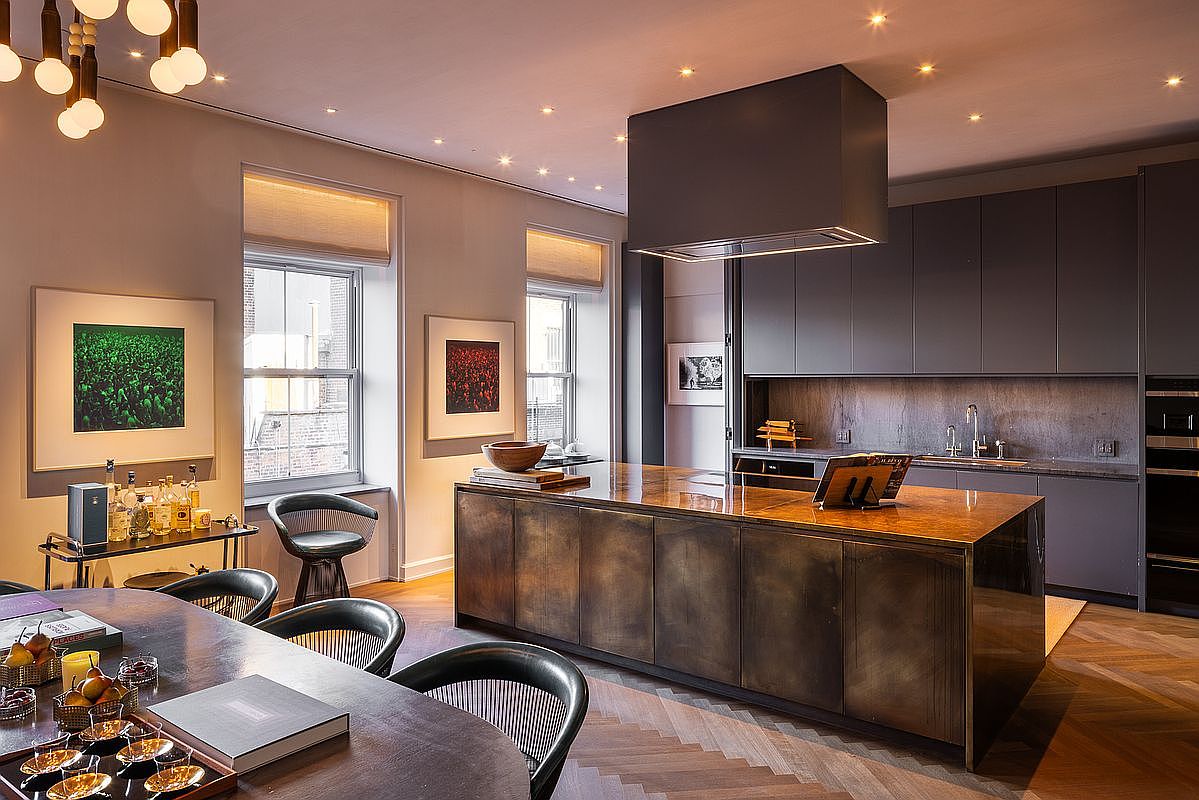 This is a modern kitchen and dining area featuring a large kitchen island with a wooden countertop and dark cabinetry. The kitchen includes sleek, gray cabinets and stainless steel appliances. The dining area features a dark wood table with modern chairs, and the room is illuminated by recessed lighting and a unique chandelier, creating a warm and inviting atmosphere.