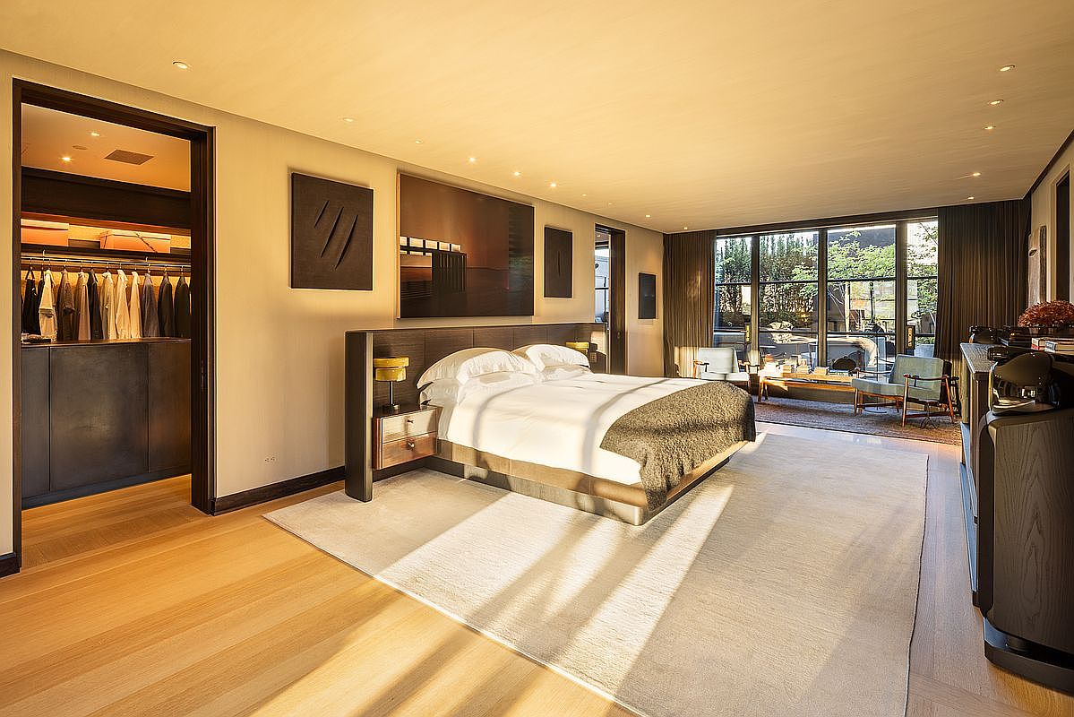 This is a spacious primary bedroom featuring a large bed with a dark wood headboard and side tables with lamps. The room has a neutral color palette with artwork on the walls and a large window offering natural light and views of the outdoors. A walk-in closet is visible through an open doorway, and the overall impression is luxurious and serene.