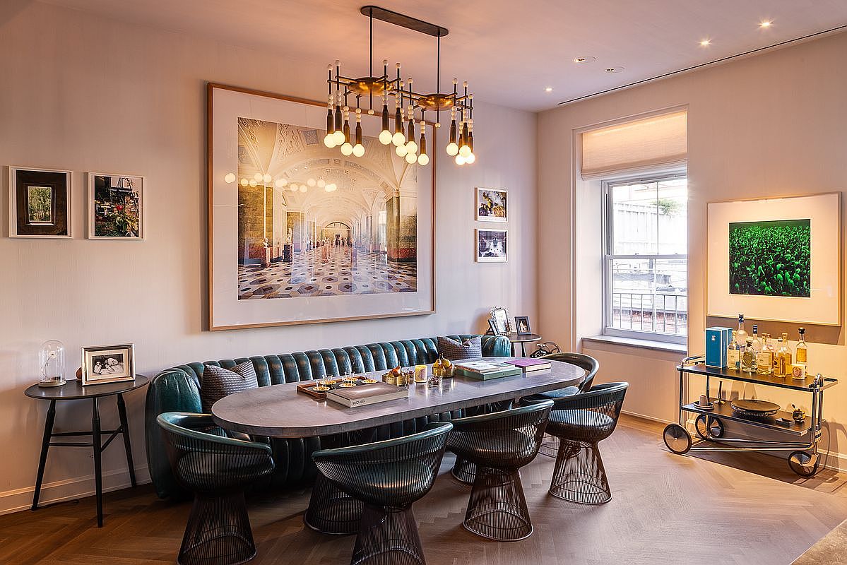 This is an interior shot of a dining room featuring a long oval table with unique wire-frame chairs and a built-in banquette. A modern chandelier hangs above the table, and large framed artwork adorns the walls, adding a sophisticated touch. A bar cart with bottles is visible near the window, suggesting an elegant space for entertaining.