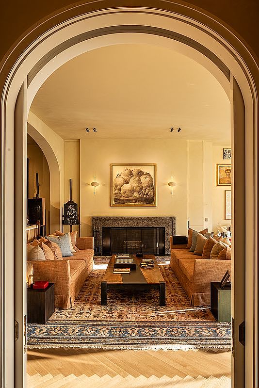 This is an interior shot of a living room, viewed through an arched doorway. The room features two sofas facing each other, a coffee table in the center, and a fireplace with a large painting above it. The decor is traditional and elegant, with a warm color palette and a patterned rug, creating a cozy and inviting atmosphere.