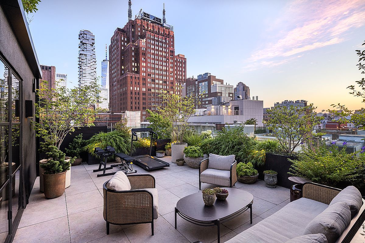 This image showcases a luxurious rooftop patio with stunning city views. The space is furnished with comfortable seating, including wicker chairs and a chaise lounge, arranged around a dark-colored coffee table. Lush greenery and potted plants add a touch of nature, while exercise equipment suggests a focus on wellness and fitness.