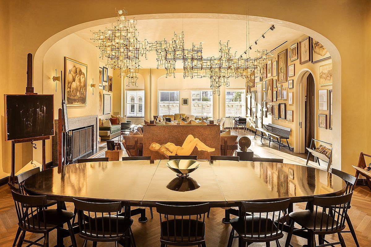 This is an interior shot of a dining room featuring a large, oval-shaped dining table surrounded by dark wooden chairs. Above the table hangs a complex, multi-tiered chandelier, and in the background, there's a glimpse into a living area with a sculpture and multiple windows. The room is decorated with numerous framed artworks on the walls, creating a sophisticated and artistic atmosphere.