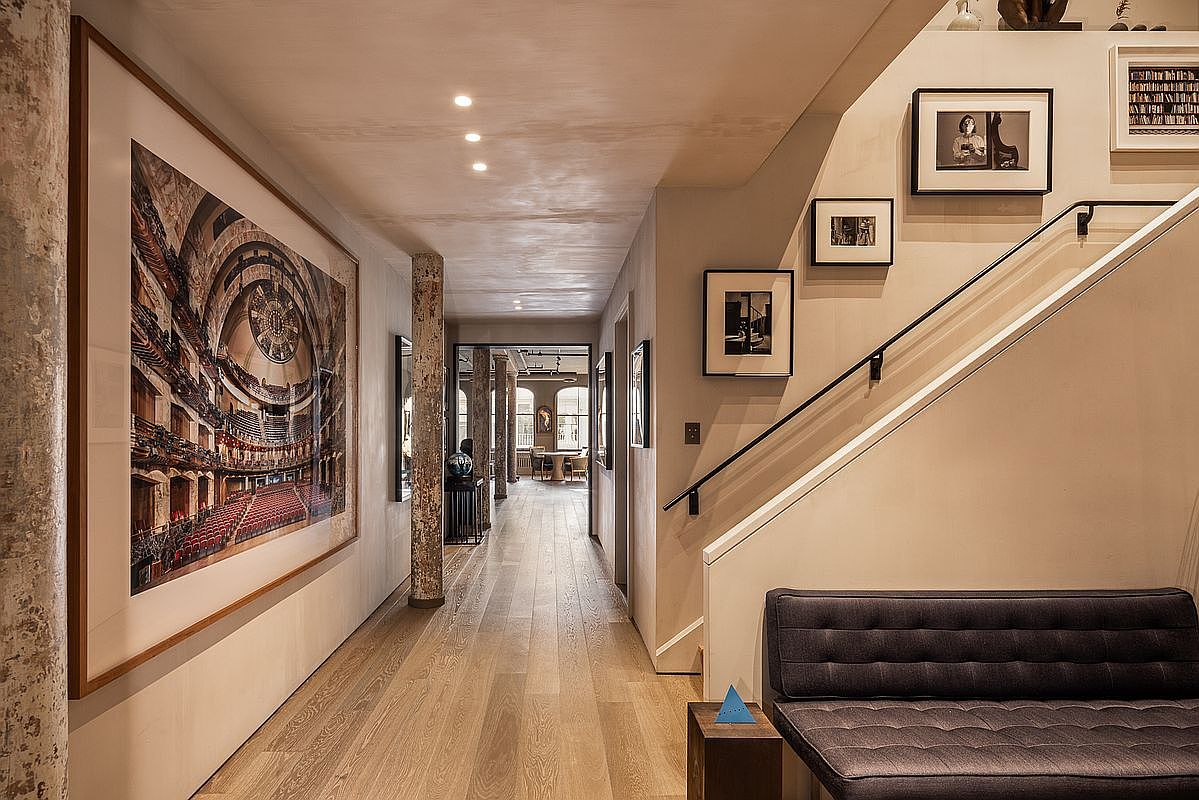This interior shot showcases a long hallway with hardwood flooring, leading to a bright, open space. A large framed artwork adorns the left wall, while a staircase with a black handrail is visible on the right, complemented by framed pictures. The hallway is well-lit with recessed lighting, creating a warm and inviting atmosphere.