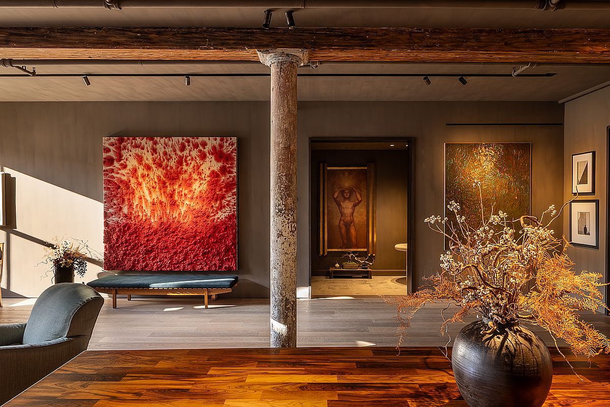 This interior shot showcases a stylish living room with a blend of modern and rustic elements. A large red and white abstract painting hangs on the wall, complemented by a dark blue bench. A decorative vase with dried branches sits on a wooden table in the foreground, while a structural column adds character to the space. The perspective is from a low angle, emphasizing the depth and artistic details of the room.