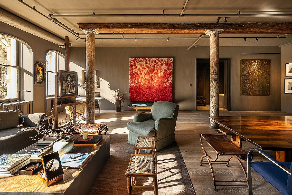 This is a spacious living room featuring exposed wooden beams and columns, adding a rustic touch to the modern interior. The room is decorated with large abstract paintings and comfortable seating arrangements, including a green armchair and a brown sofa. Natural light floods the space through large arched windows, highlighting the hardwood floors and creating a warm, inviting atmosphere.