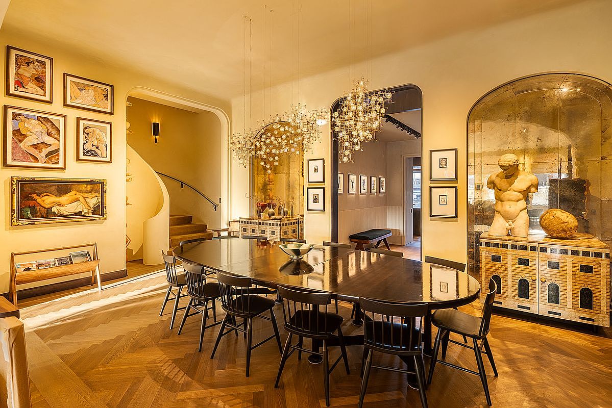 This is an elegant dining room featuring a long, dark wood dining table surrounded by black chairs, illuminated by two modern chandeliers. The room boasts hardwood floors in a herringbone pattern and is decorated with artwork and a sculpture displayed on a cabinet with a mirrored backdrop. The space exudes sophistication and is perfect for formal dining and entertaining.