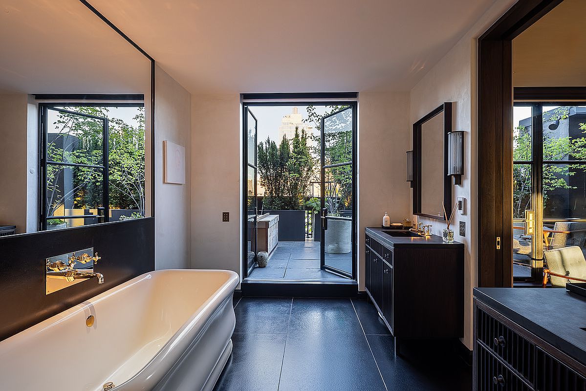 This is a luxurious primary bathroom featuring a freestanding bathtub with gold fixtures, a dark wood vanity with a vessel sink, and black tile flooring. French doors open to a private balcony with greenery, creating a spa-like atmosphere. The space is well-lit and exudes a modern, sophisticated style.